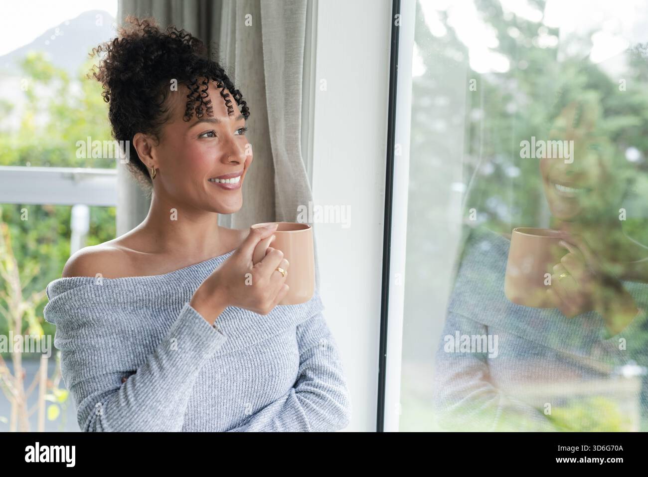 Frau in ihren 30ern, die zu Hause am Fenster stand und Keramikbecher hielt und einen Schulterpullover trug Stockfoto