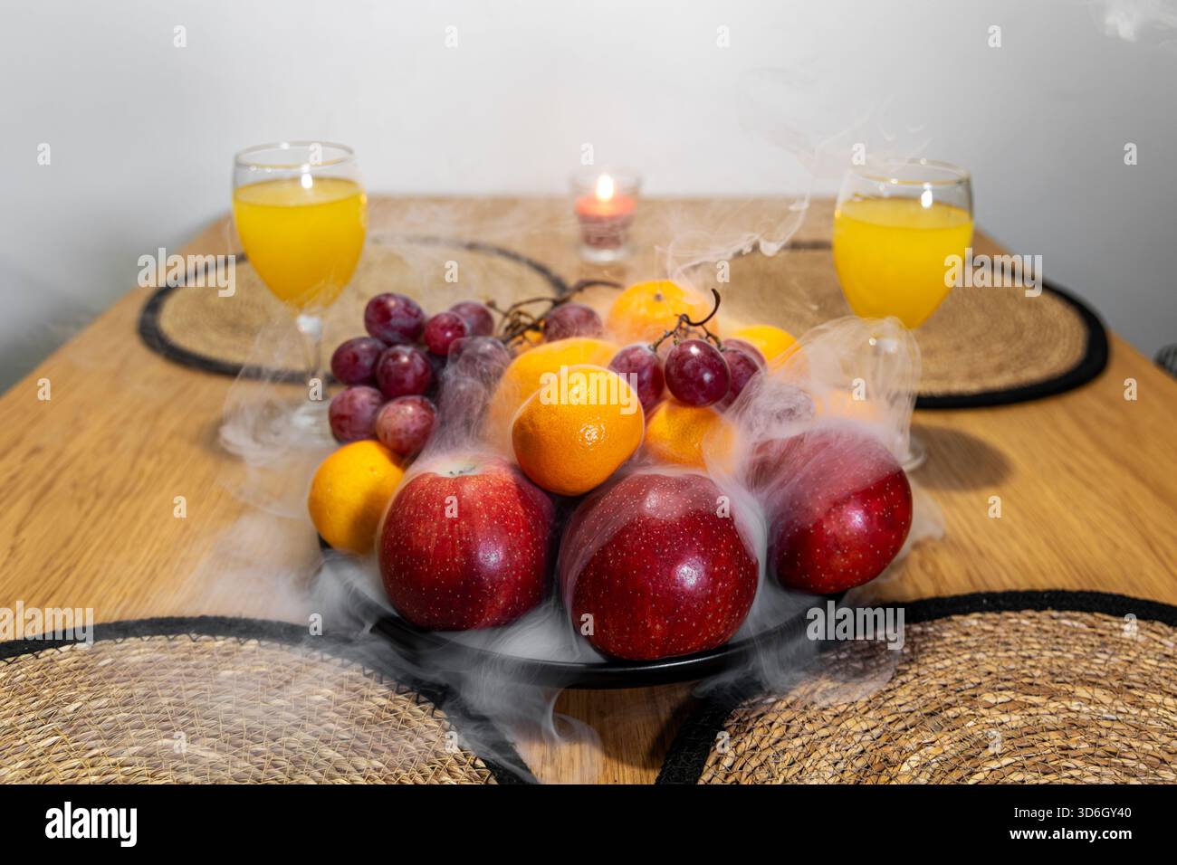 Obstplatte mit Raucheffekt auf Esstisch. Künstlerische Darstellung von frischem Obst mit Raucheffekt auf einem hölzernen Esstisch, flankiert von Orange Stockfoto