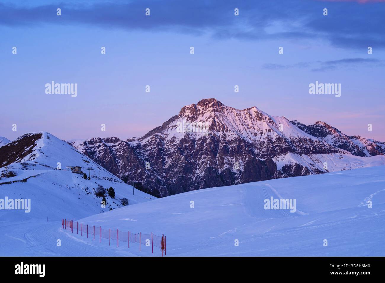 Piani di Bobbio, Italien - 24.12.2022: Schneebedeckter Berggipfel leuchtet im schwindenden Licht des Sonnenuntergangs. Im Vordergrund stehen schneebedeckte Pisten und Skistöcke, o Stockfoto