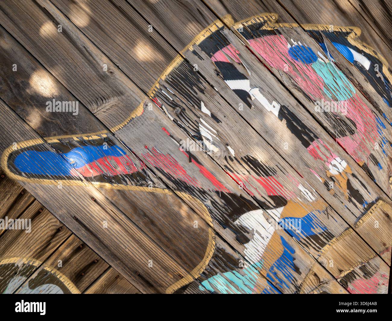 Nahaufnahme eines verwitterten, handbemalten Entendesigns auf diagonalen Holzdielen. Schichten abblätternder Farbe in Rot, Blau, Petrol, Schwarz, weiß, und Gold. Stockfoto