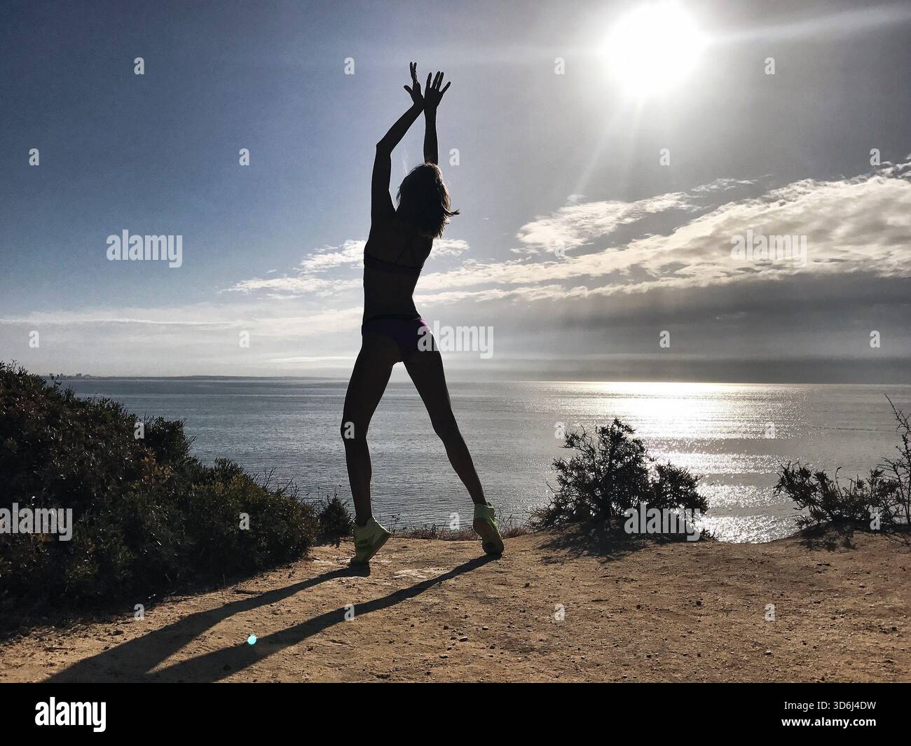 Silhouette einer Frau in Tänzerin-Pose mit Flügeln erhobenen Armen, die über der Küste gegen die Sonne stehen. Weibliche Figur vor dem Meer. Stockfoto