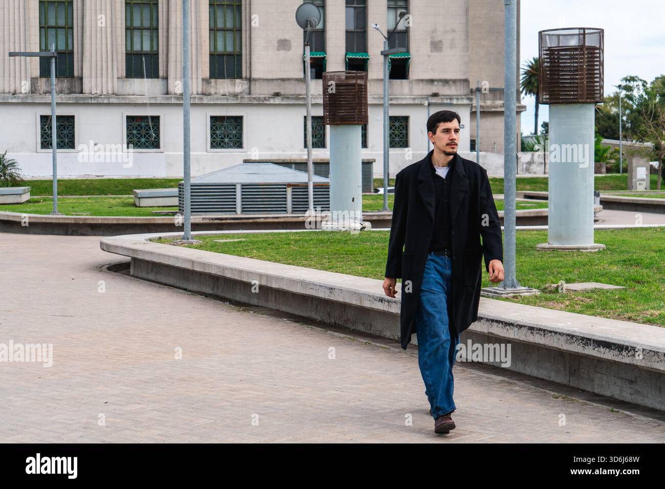 Junger Mann in schwarzem Mantel und Jeans spaziert nachdenklich durch einen urbanen Park neben moderner Architektur, einsam und introspektiv Stockfoto