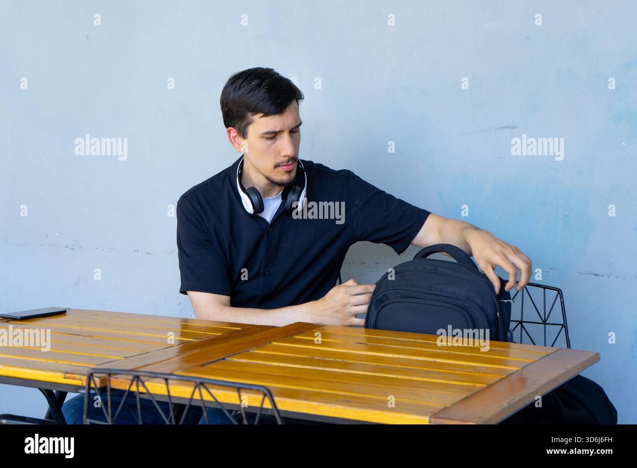 Junger Mann mit Kopfhörern um den Hals, der im Café im Freien sitzt und den Rucksack öffnet, um einen Gegenstand daraus zu nehmen Stockfoto
