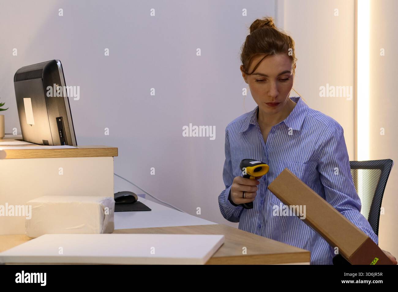 Bediener in blauem Hemd scannt vertikale Kartonverpackungen mit Scanner neben Monitor und Maus. Stockfoto