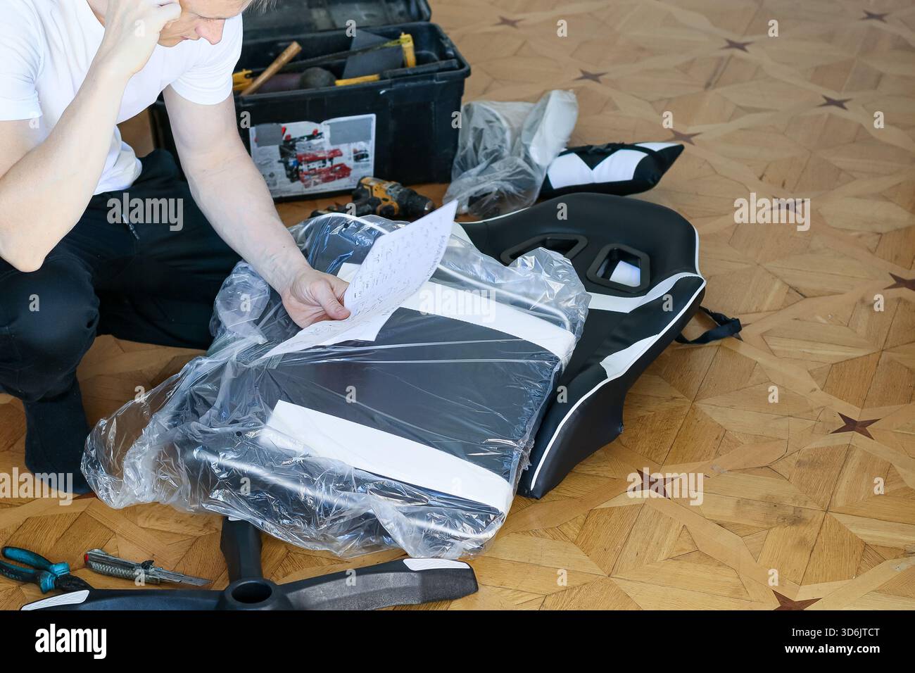 Ein Mann in lässiger Kleidung kniet auf Holzboden, verwirrter Ausdruck, während er den Entwurf für die Zusammenstellung ergonomischer Spielstühle mit Werkzeugen und Packa studiert Stockfoto