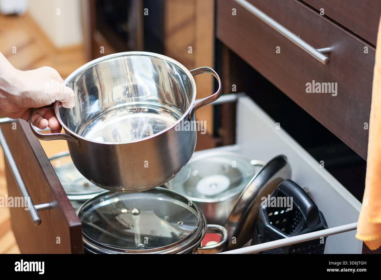 Nahaufnahme einer Hand, die eine hölzerne Küchenschublade öffnet, gefüllt mit organisiertem Edelstahl-Kochgeschirr, Töpfen, Pfannen und Deckeln in einem modernen Innenraum. Stockfoto