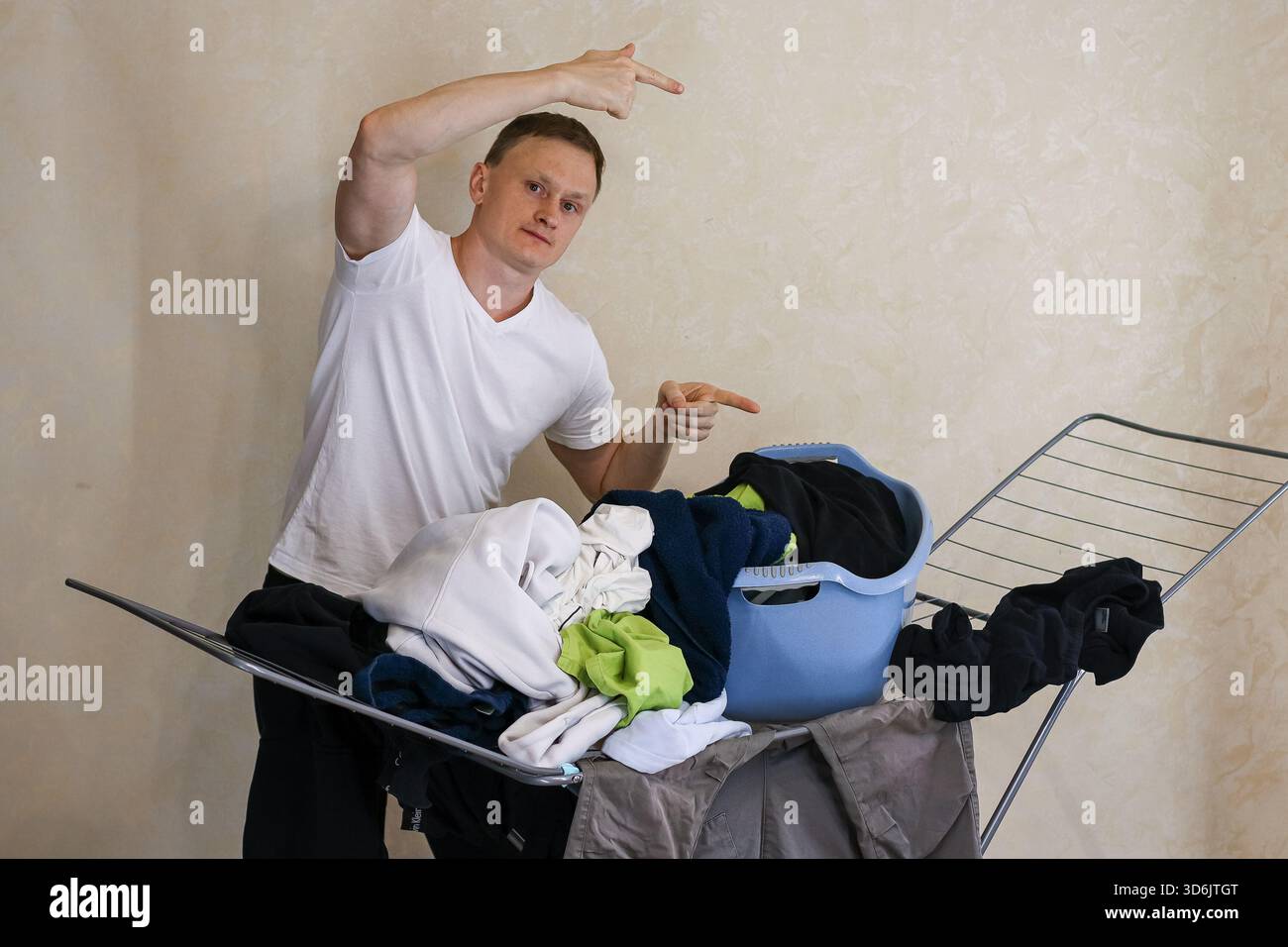 Ein junger Mann in weißem T-Shirt bewegt sich auf einen Haufen bunter Kleidung auf einem Metalltrockenständer mit blauem Korb in der Nähe, Hauswäscherei Szene Stockfoto