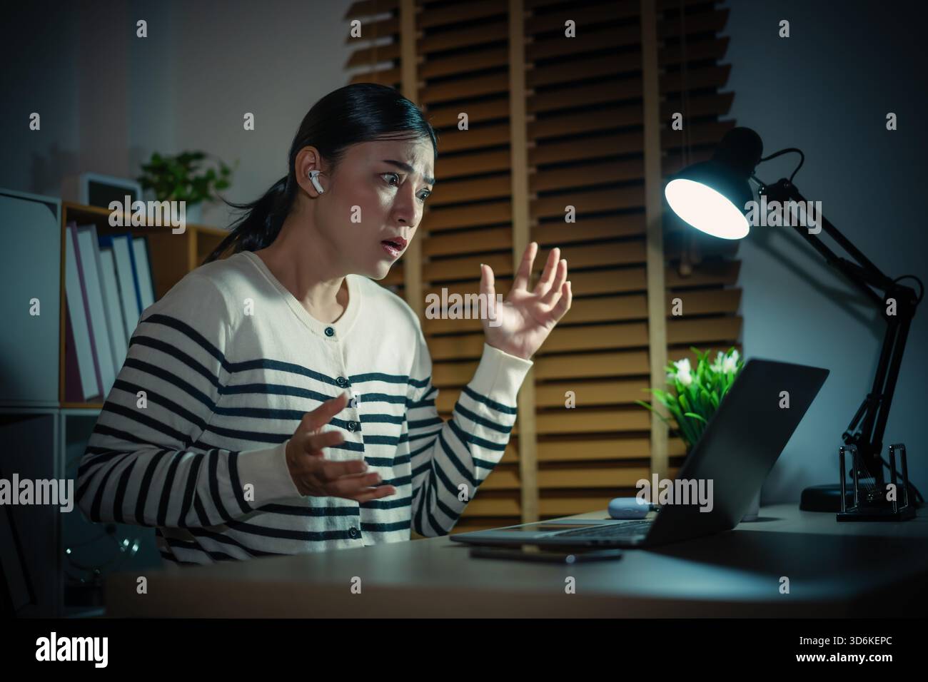 Stressige Frau mit Laptop und schnurlosen Ohrstöpseln, die nachts Videoanrufe tätigen Stockfoto