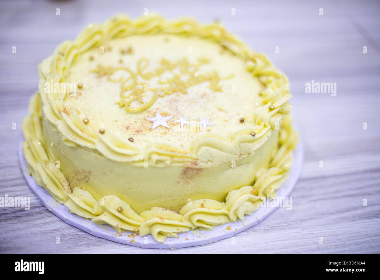Eine gelbe Milchkuche, dekoriert mit Paspeln an Buttercremeborten, goldenen Perlen, essbarem Glitzer und weißen Sternakzenten, platziert auf einer Holzoberfläche Stockfoto