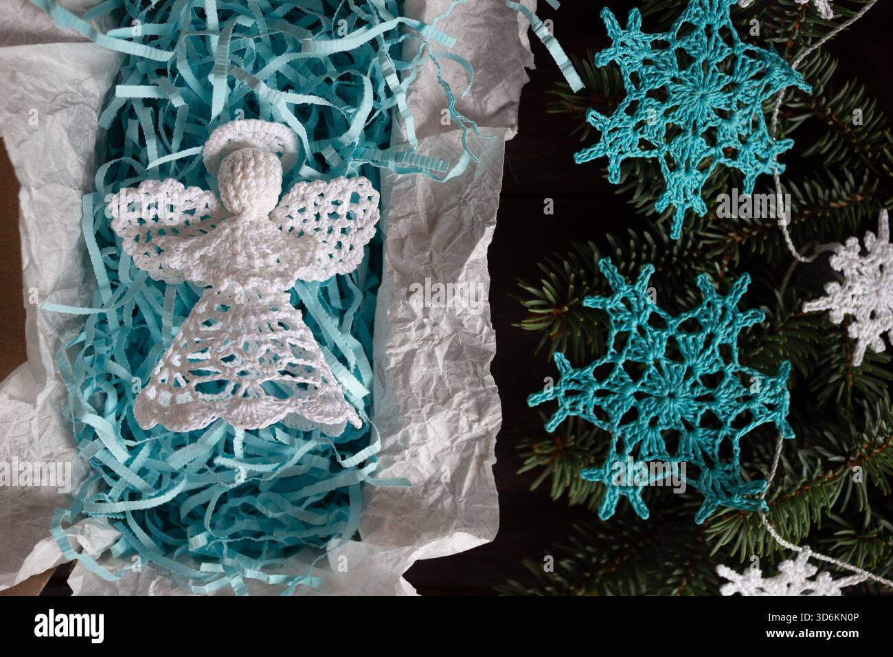 Handgefertigter weißer Weihnachtsengel in Geschenkverpackung mit blauem Schredderpapier, umgeben von gehäkelten Schneeflocken-Ornamenten und Tannenzweigen, Top Stockfoto