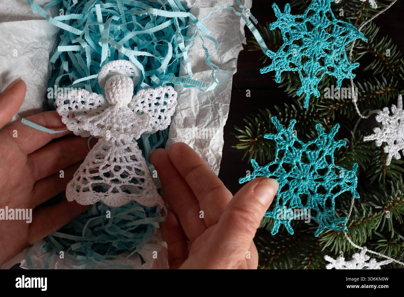 Handgefertigter weißer Weihnachtsengel in Geschenkverpackung mit blauem Schredderpapier, umgeben von gehäkelten Schneeflocken-Ornamenten und Tannenzweigen, Top Stockfoto