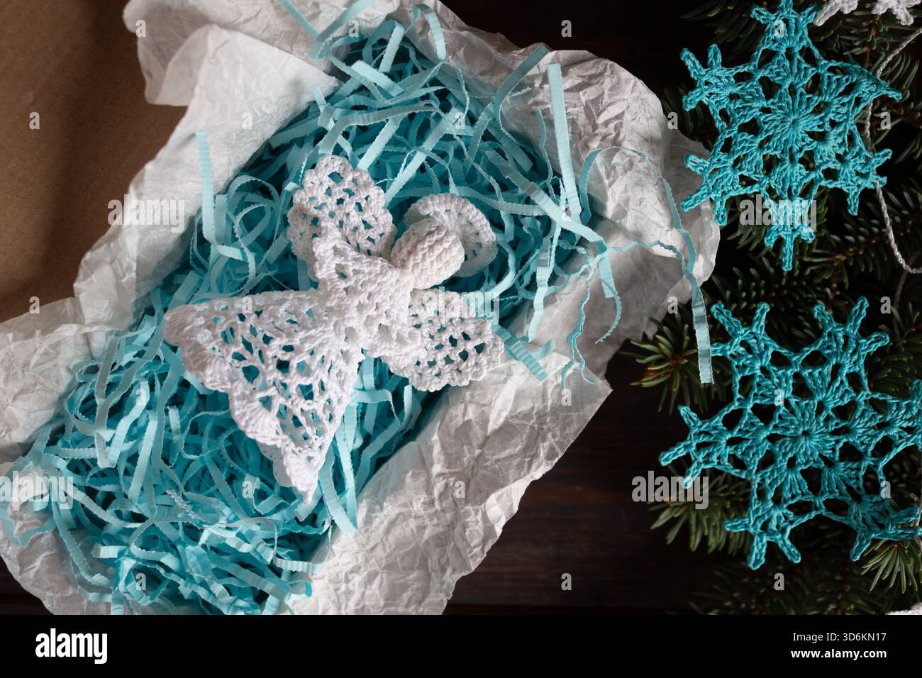 Handgefertigter weißer Weihnachtsengel in Geschenkverpackung mit blauem Schredderpapier, umgeben von gehäkelten Schneeflocken-Ornamenten und Tannenzweigen, Top Stockfoto