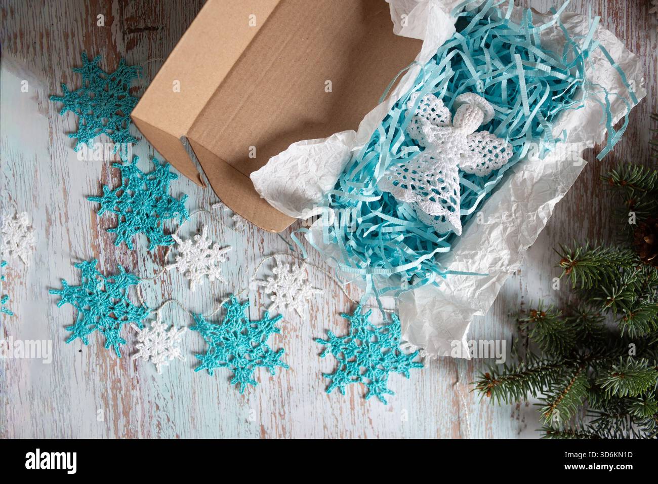 Handgefertigter weißer Weihnachtsengel in Geschenkverpackung mit blauem Schredderpapier, umgeben von gehäkelten Schneeflocken-Ornamenten und Tannenzweigen, Top Stockfoto