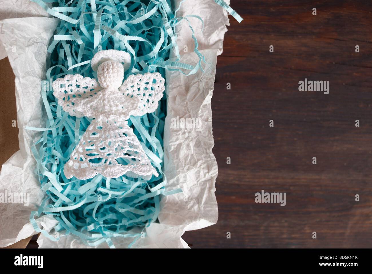 Handgefertigter weißer Weihnachtsengel in Geschenkverpackung mit blauem Schredderpapier, umgeben von gehäkelten Schneeflocken-Ornamenten und Tannenzweigen, Top Stockfoto