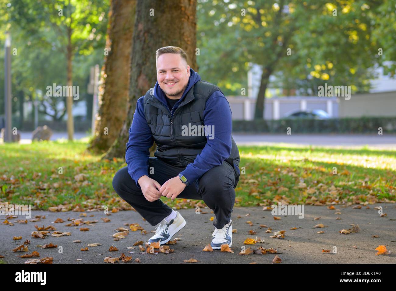 Junger Mann hockt auf einem Parkweg, umgeben von Herbstlaub, und lächelt warm in die Kamera. Stockfoto