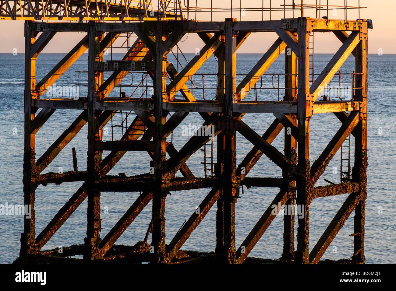 Verwitterter hölzerner Pilotensteg, beleuchtet durch warmes Sonnenlicht mit ruhigem Meer und sanftem Horizont dahinter Stockfoto