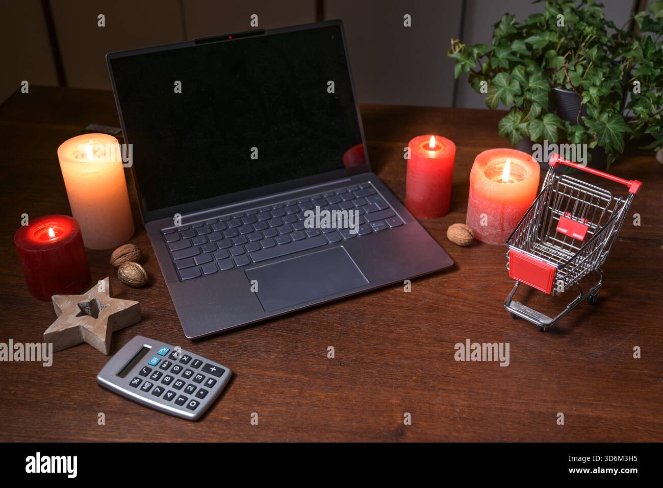 Laptop, Taschenrechner und ein kleiner Einkaufswagen umgeben von Kerzen und Weihnachtsdekoration auf einem dunklen hölzernen Bürotisch, Online-Shopping-Budget und h Stockfoto