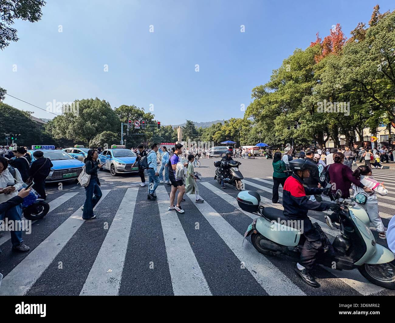 Fußgänger navigieren an einer Straßenkreuzung in einer chinesischen Stadt auf einem geschäftigen Zebraweg, an dem Autos und Motorräder warten und vorbeifahren. Repräsentiert URBAN Stockfoto