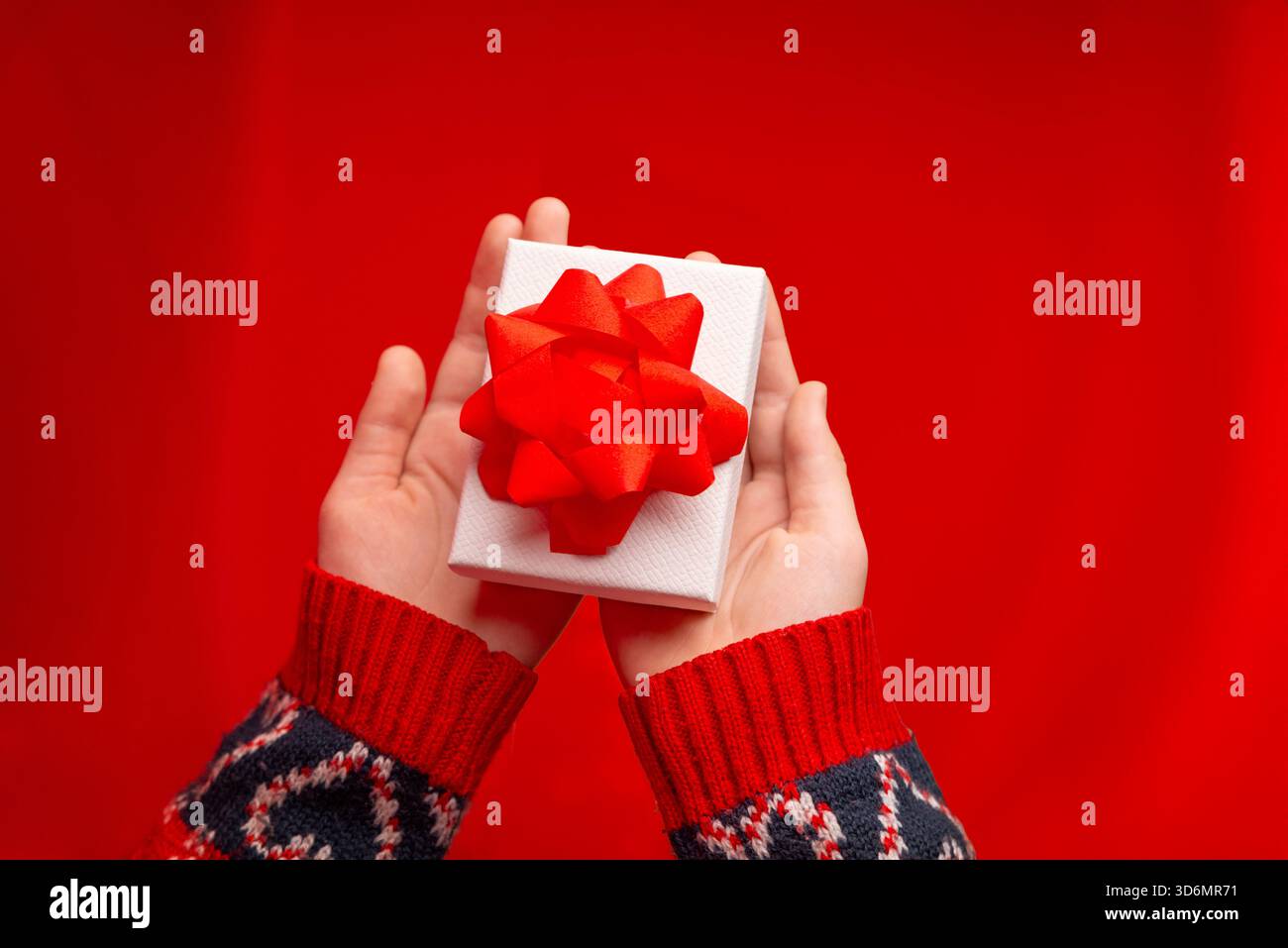 Blick von oben auf die Hände, die eine weiße Geschenkbox mit roter Schleife auf hellrotem Hintergrund halten, perfekt für festliche Kampagnen und Weihnachtskreativen Stockfoto