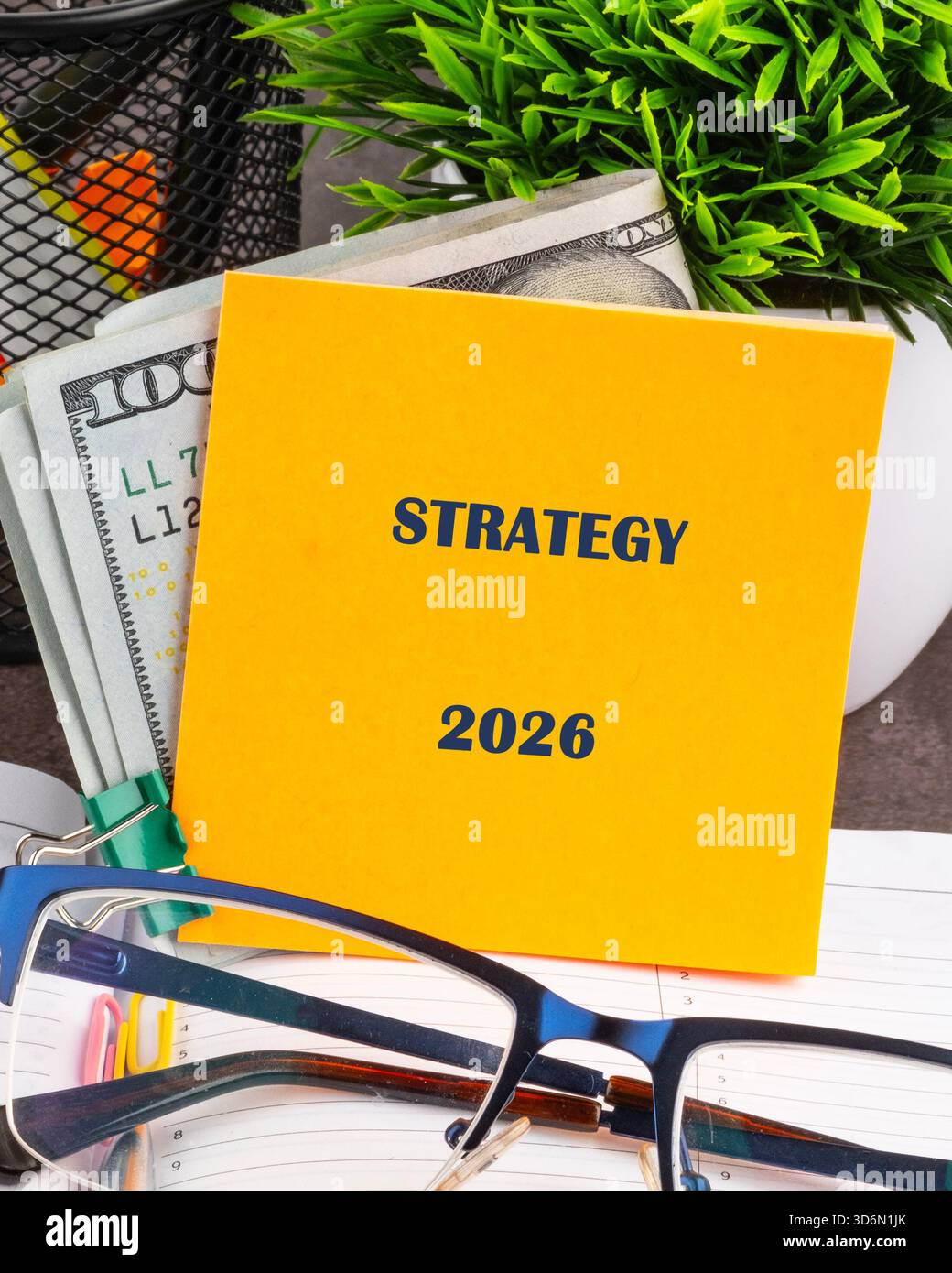 Ein gelber Haftnotiz mit der Aufschrift Strategie 2026 befindet sich auf einem Notebook mit Geld. Eine Brille befindet sich im Vordergrund, und eine kleine grüne Pflanze verleiht ihr einen Hauch Stockfoto
