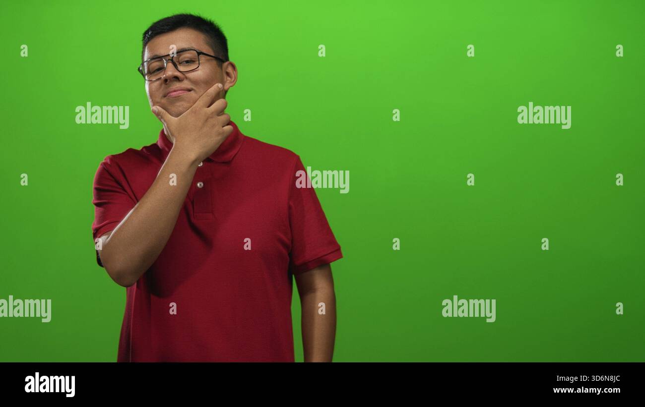 Junger Mann mit Hand auf dem Kinn, der in grünem Hintergrund im Studio posiert, mit rotem Polo und Brille; nachdenkliche Reflexion. Stockfoto
