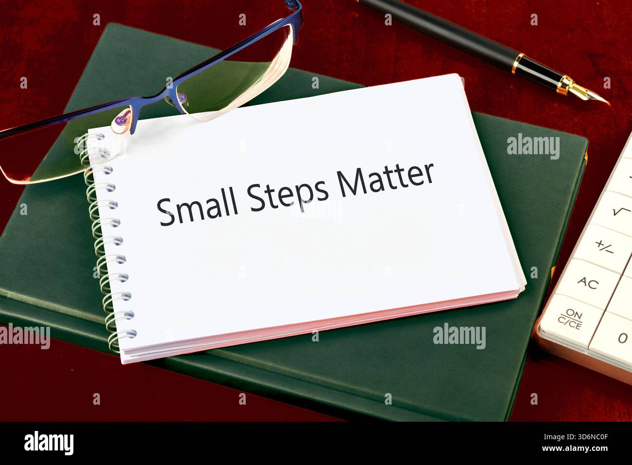 Eine organisierte Szene auf einem hölzernen Schreibtisch mit einem Notizbuch mit kleinen Schritten auf einem grünen Buch, begleitet von einer Brille, einem Stift und einer Kalkulation Stockfoto