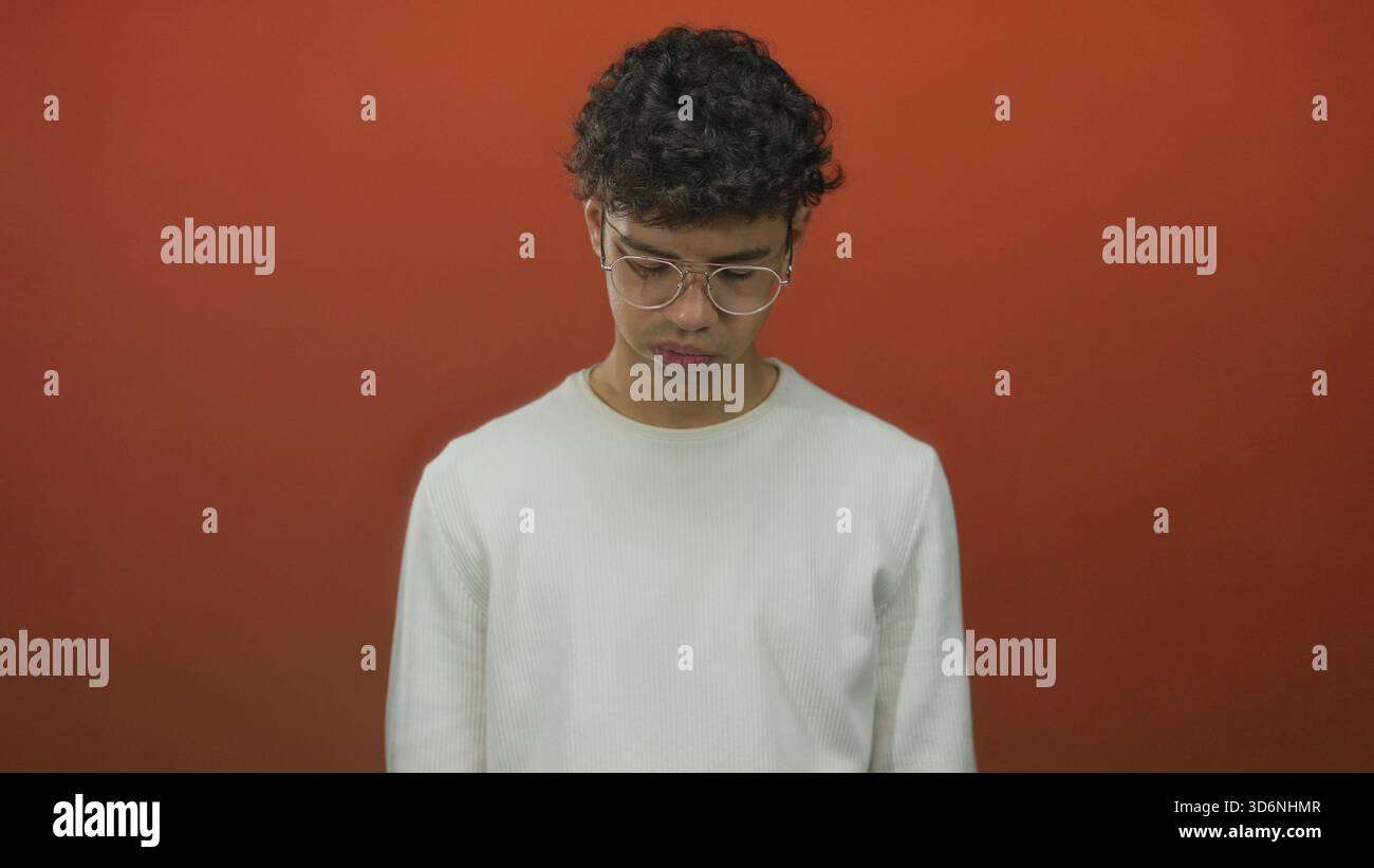 Mann mit Brille, lockigen Haaren und weißem Hemd schaut mit gebeugter Haltung gegen die orangene Studiowand; Nachsicht. Stockfoto