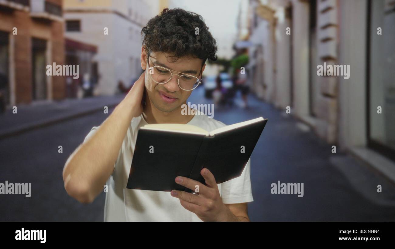 Mann liest Buch mit Hand am Kinn, trägt Brille und weißes T-Shirt auf der sonnenbeschienenen Straße in der Stadt; Neugier-Reflexion. Stockfoto