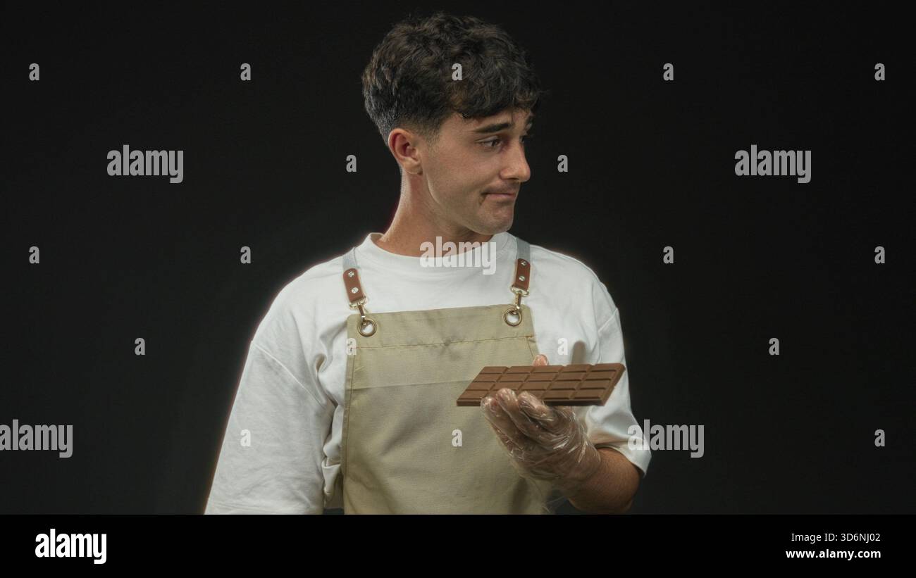 Junger hispanischer Mann, der mit Handschuhen eine Schokoladentafel vor schwarzem Hintergrund hält; ruhige Enttäuschung. Stockfoto