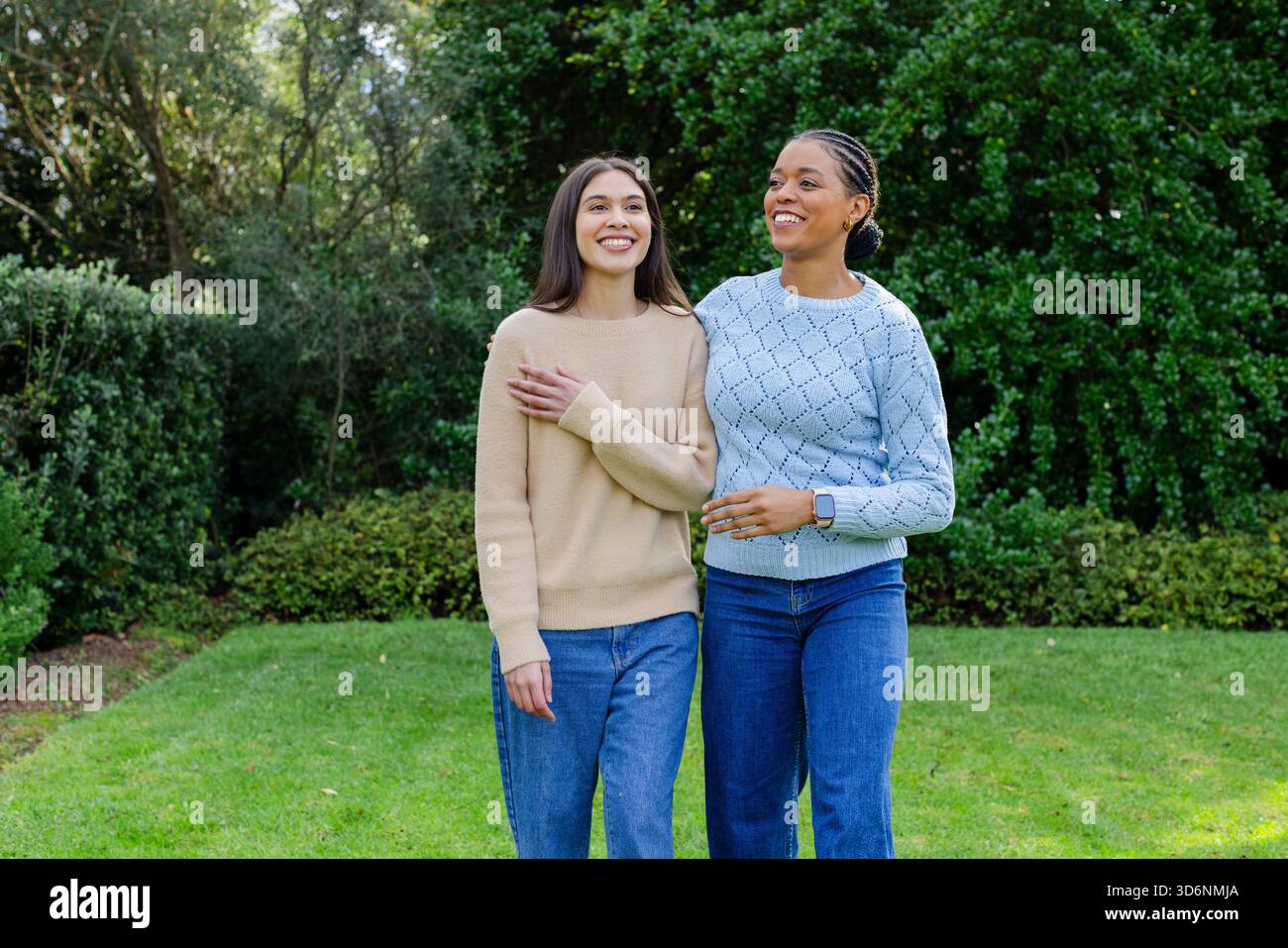 Weibliche Freunde, die Arm in Arm auf dem Rasen laufen, tragen lässige Pullover und Jeans im Park Stockfoto