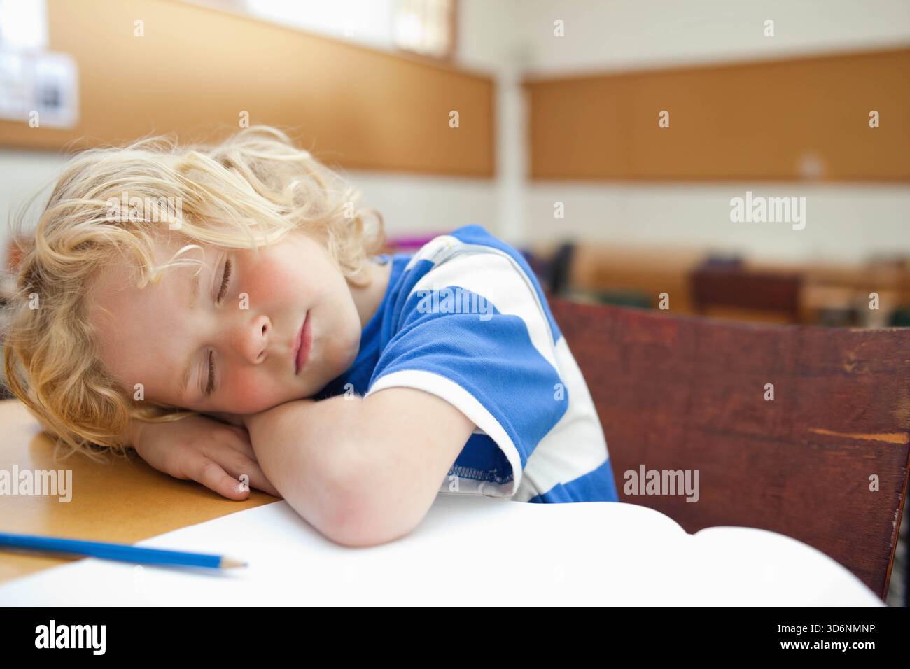 Junge, der auf gefalteten Armen auf einem Holzschreibtisch in der Nähe eines offenen Notizbuchs schläft, blauer Bleistift im Klassenzimmer. Nickerchen, Pupille, Tisch, Tageslicht, Wärme, Bokeh, ruhig Stockfoto
