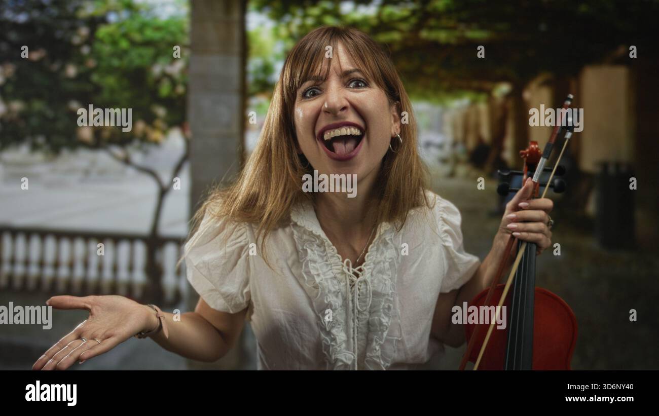 Die Frau winkt die Hand, während sie Geige und Bogen am Hals hält, auf einer von Bäumen gesäumten Straße neben einem Steingebäude; spielerische Freude. Stockfoto