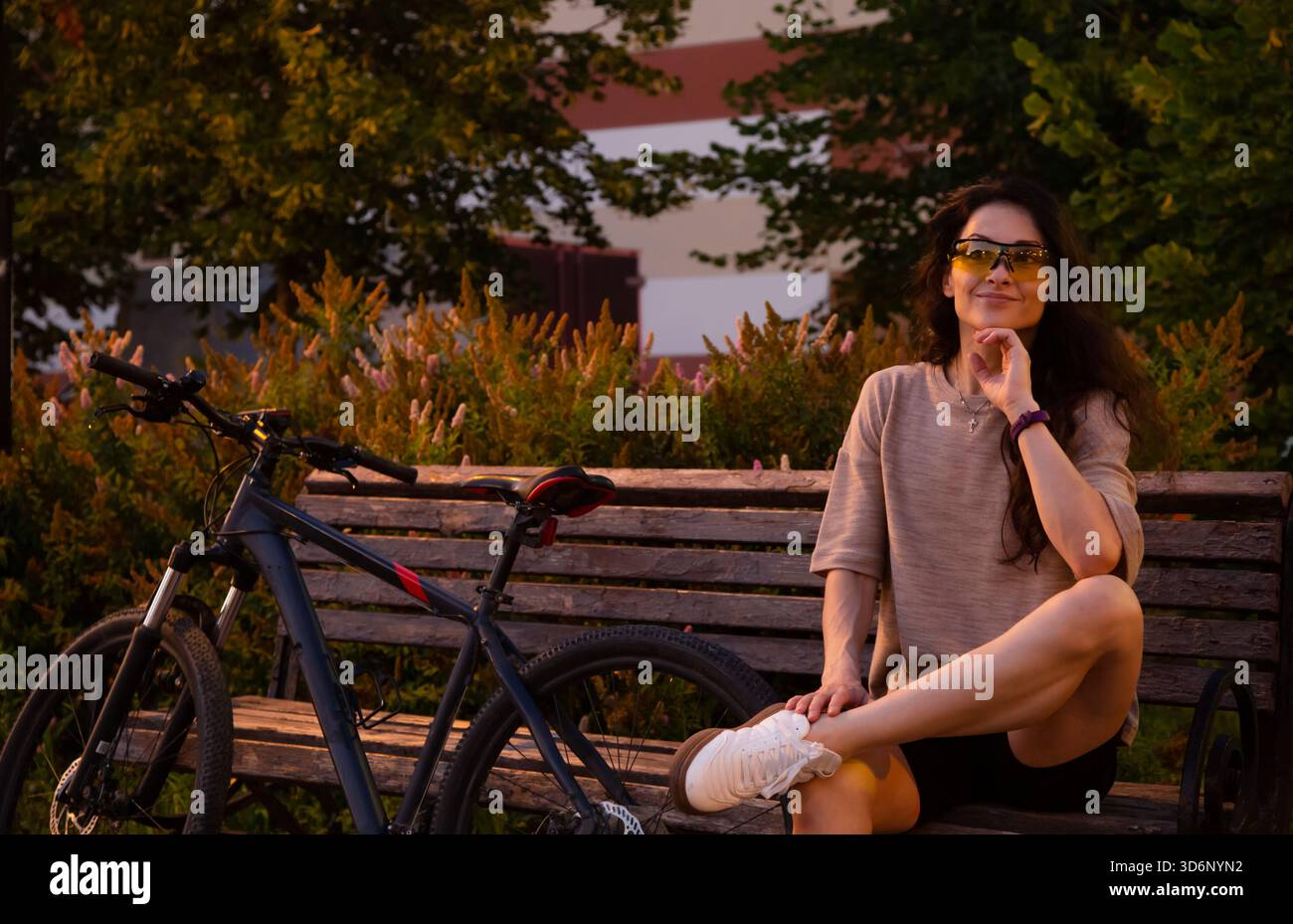 Entspannende denken Sie Frau, die mit ihrem Fahrrad auf der Bank sitzt und in einer Sportschutzbrille in einem Stadtpark im Abendlicht von Straßenlaternen schaut. Person Stockfoto