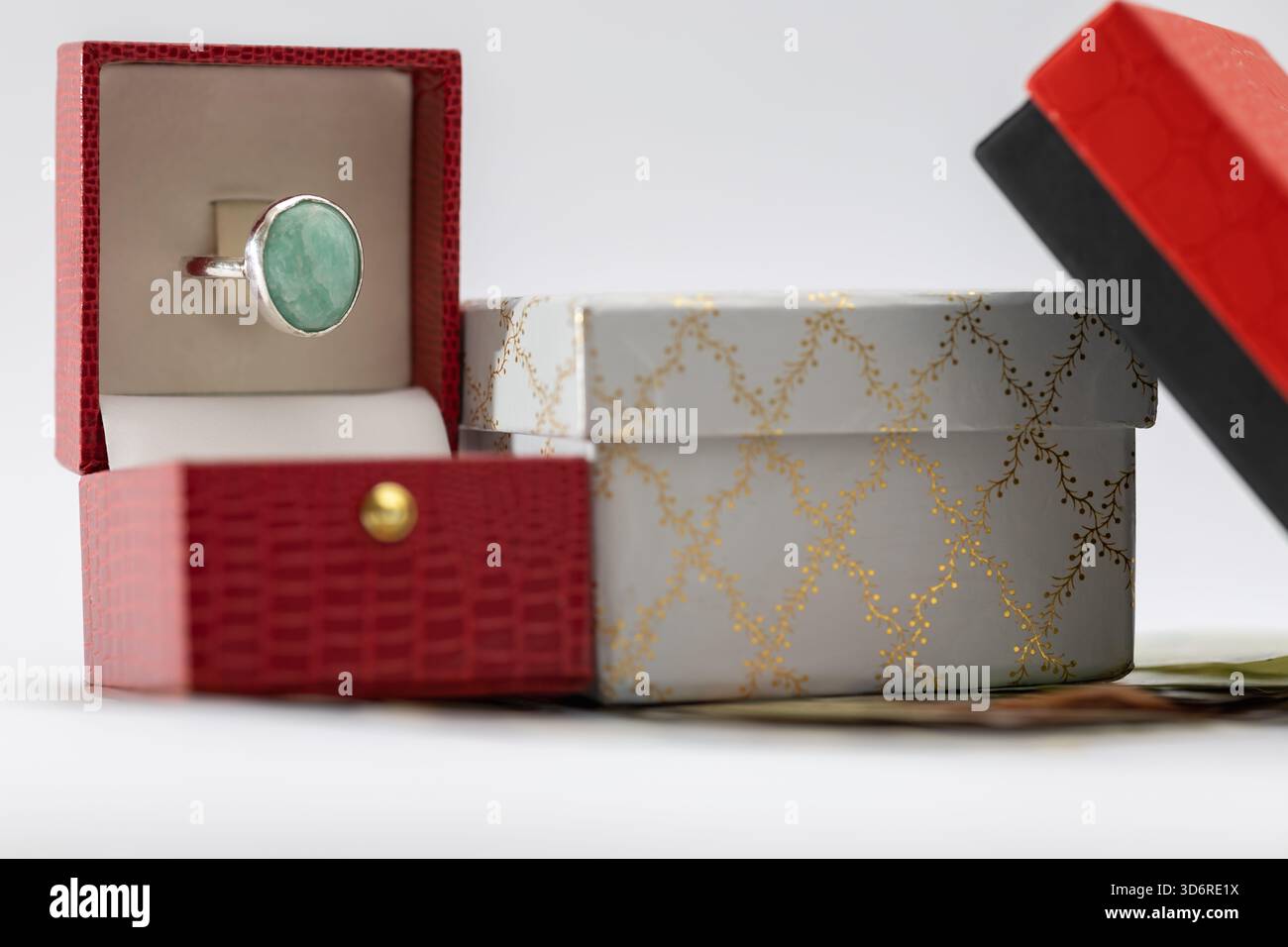 Ein eleganter Silberring mit einem großen Aquamarin-/Smaragdstein in einer offenen roten Geschenkbox, neben einer dekorativen, weiß-goldenen Mustergarnitur und einem schwarzen bo Stockfoto