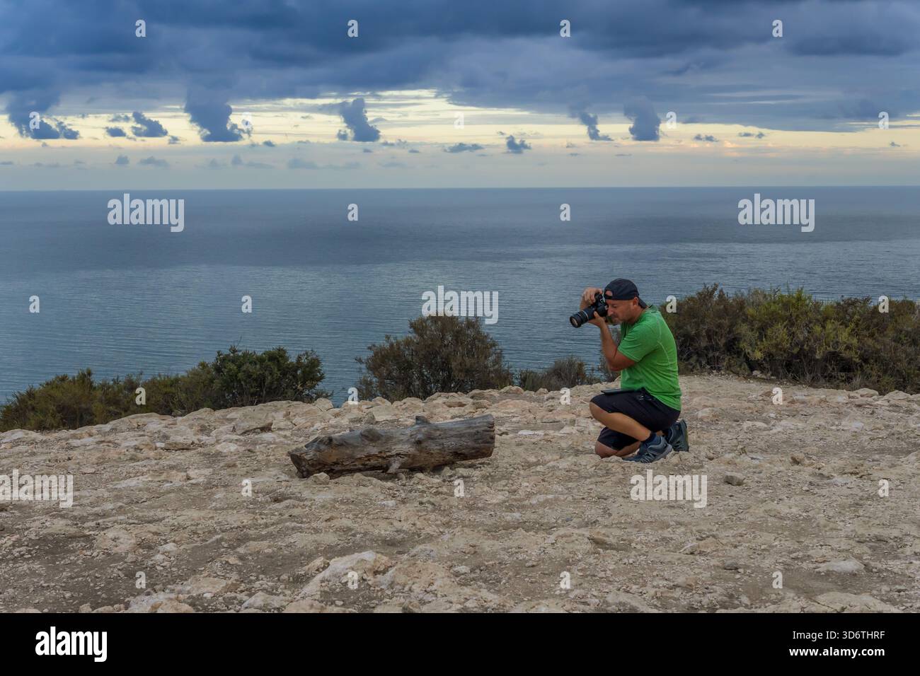 Fotograf macht eine Nahaufnahme eines hölzernen Zweigs auf dem Boden mit einem verschwommenen Wasser in der Stadtbucht und einem bewölkten Abendhimmel im Hintergrund. Stockfoto