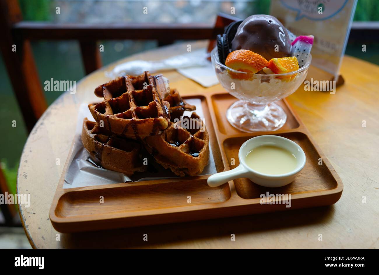 Eine köstliche Dessertkombination auf einem Holztisch, einschließlich Waffeln, Kondensmilch und einer Schaufel Schokoladeneis mit Fruchtstücken Stockfoto