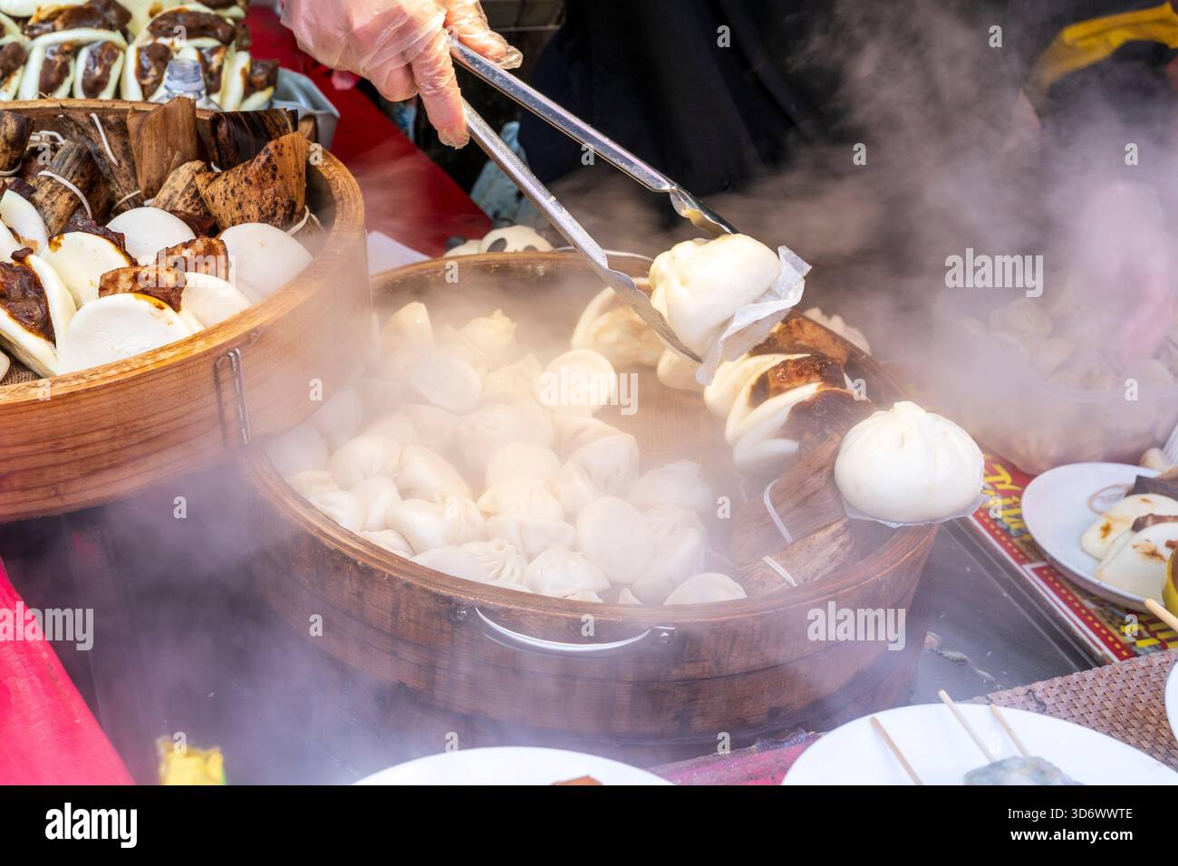 Nahaufnahme einer Hand, die eine Zange hält, die einen Teig aus einer Bambusschüssel mit Teigtaschen und gedämpften Brötchen an einem heißen Imbissstand in Chinatown entfernt Stockfoto