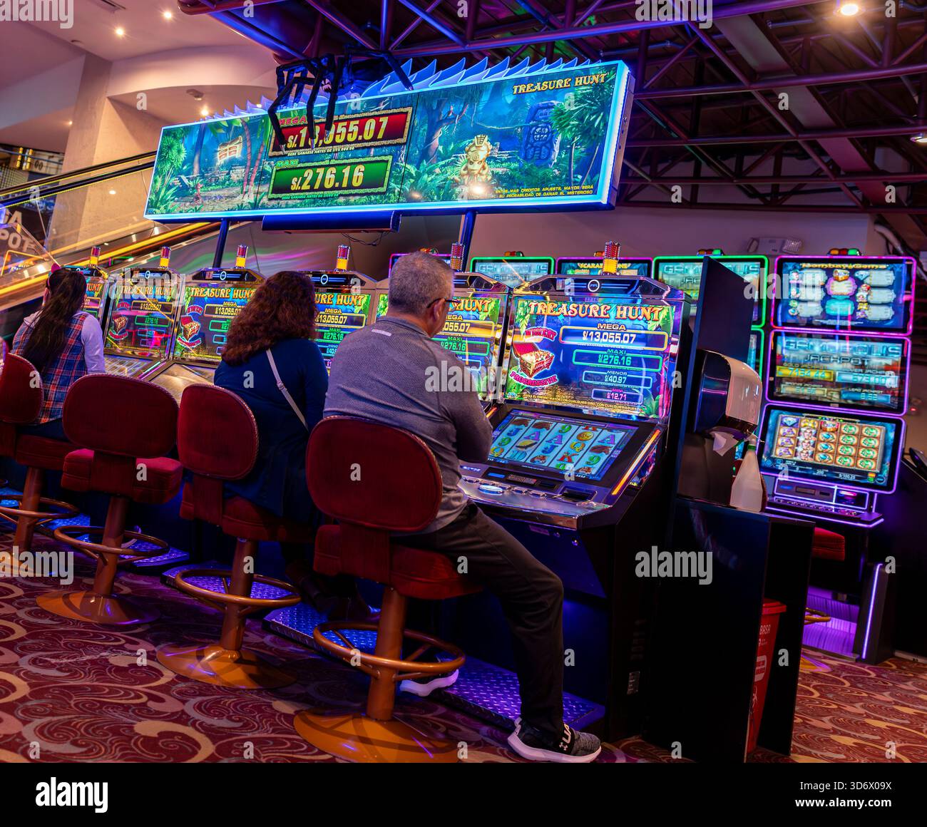 Spieler sitzen an den bunten Treasure Hunt Spielautomaten in einem hellen Casino mit beleuchteten Bildschirmen und Spiellichtern. Stockfoto