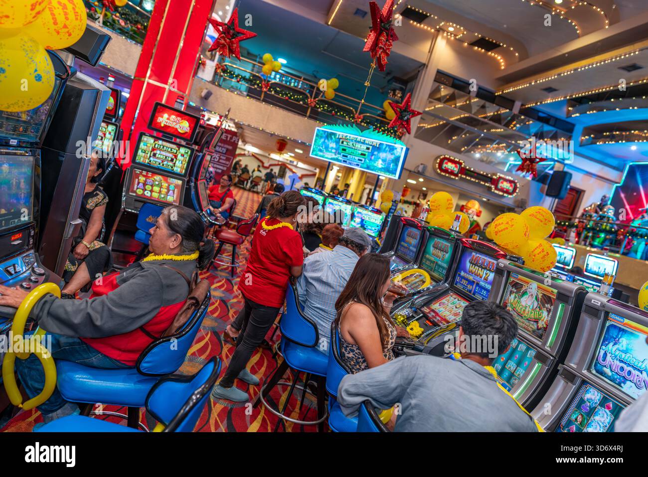 In einem festlichen Casino mit hellen Lichtern, bunten Bildschirmen, Ballons und einer lebhaften Unterhaltungsatmosphäre spielen die Leute Spielautomaten. Stockfoto
