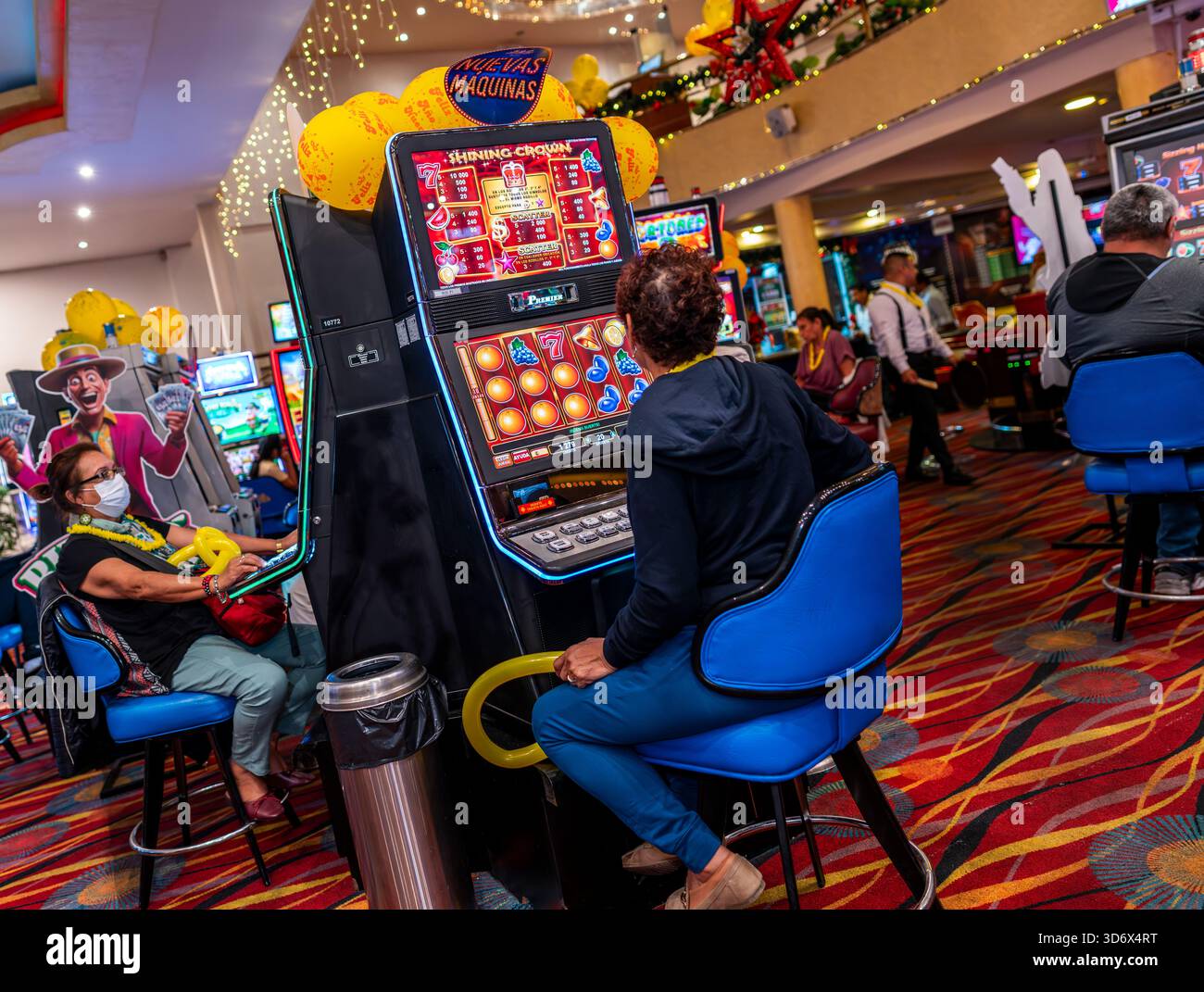 In einem festlichen Kasino mit hellen Lichtern, bunten Bildschirmen, Ballons und einer lebhaften Unterhaltungsatmosphäre spielen die Leute Spielautomaten. Stockfoto