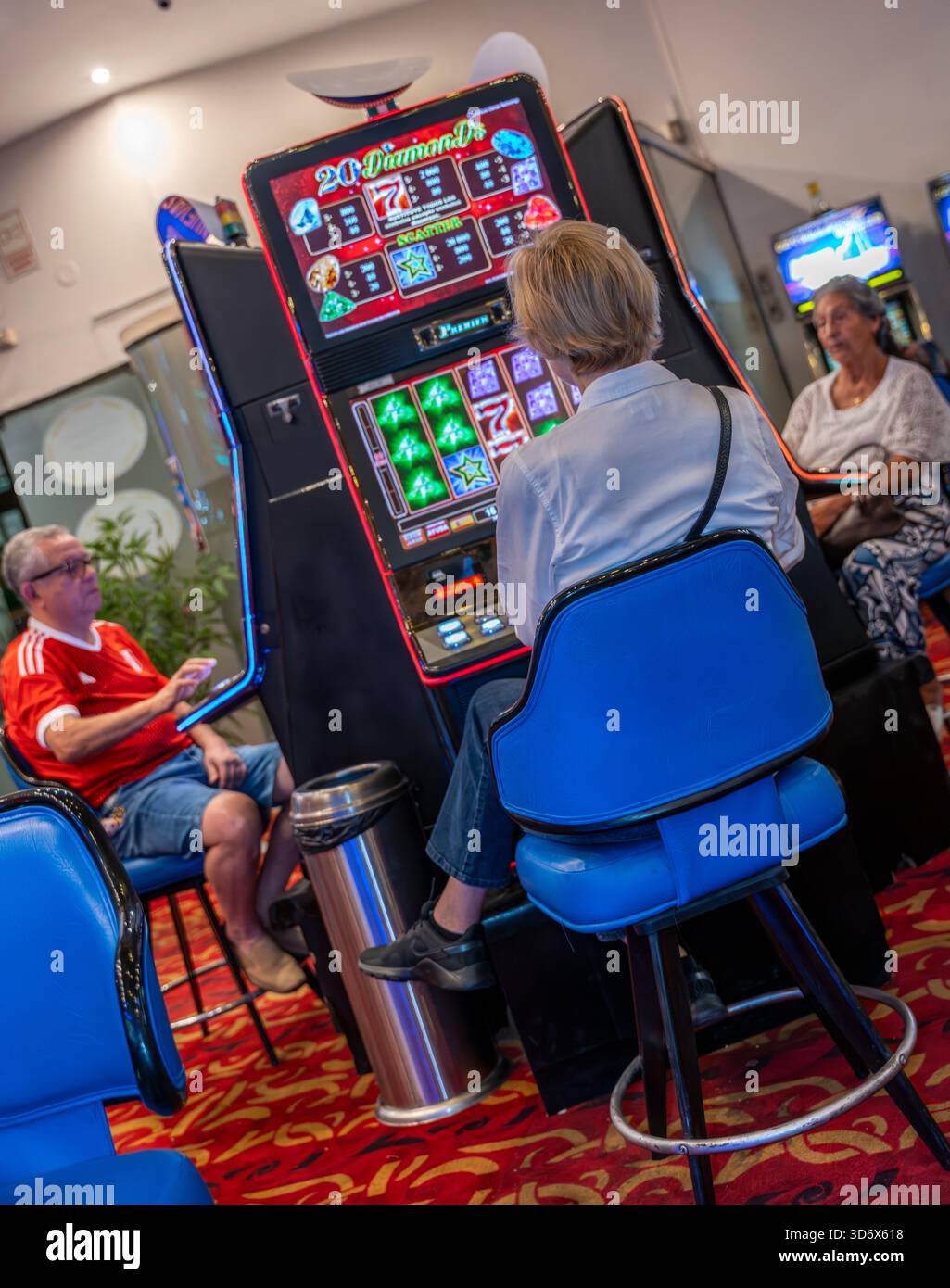 In einem Kasino mit rotem Teppich, blauen Stühlen und einer entspannten Spielumgebung saßen die Leute an einem 20-Diamonds-Spielautomaten. Stockfoto