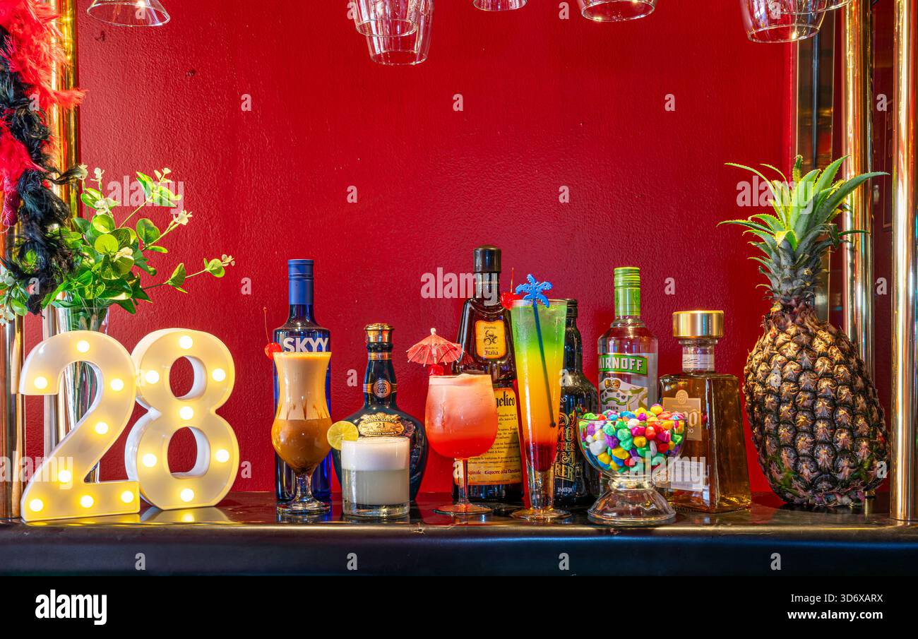 Farbenfrohe Cocktails, Spirituosenflaschen, Schüssel und dekorative Elemente auf einem festlichen Bartisch mit leuchtend roter Hintergrundbeleuchtung. Stockfoto