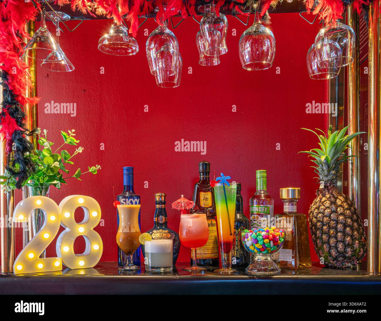 Farbenfrohe Cocktails, Spirituosenflaschen, Schüssel und dekorative Elemente auf einem festlichen Bartisch mit leuchtend roter Hintergrundbeleuchtung. Stockfoto