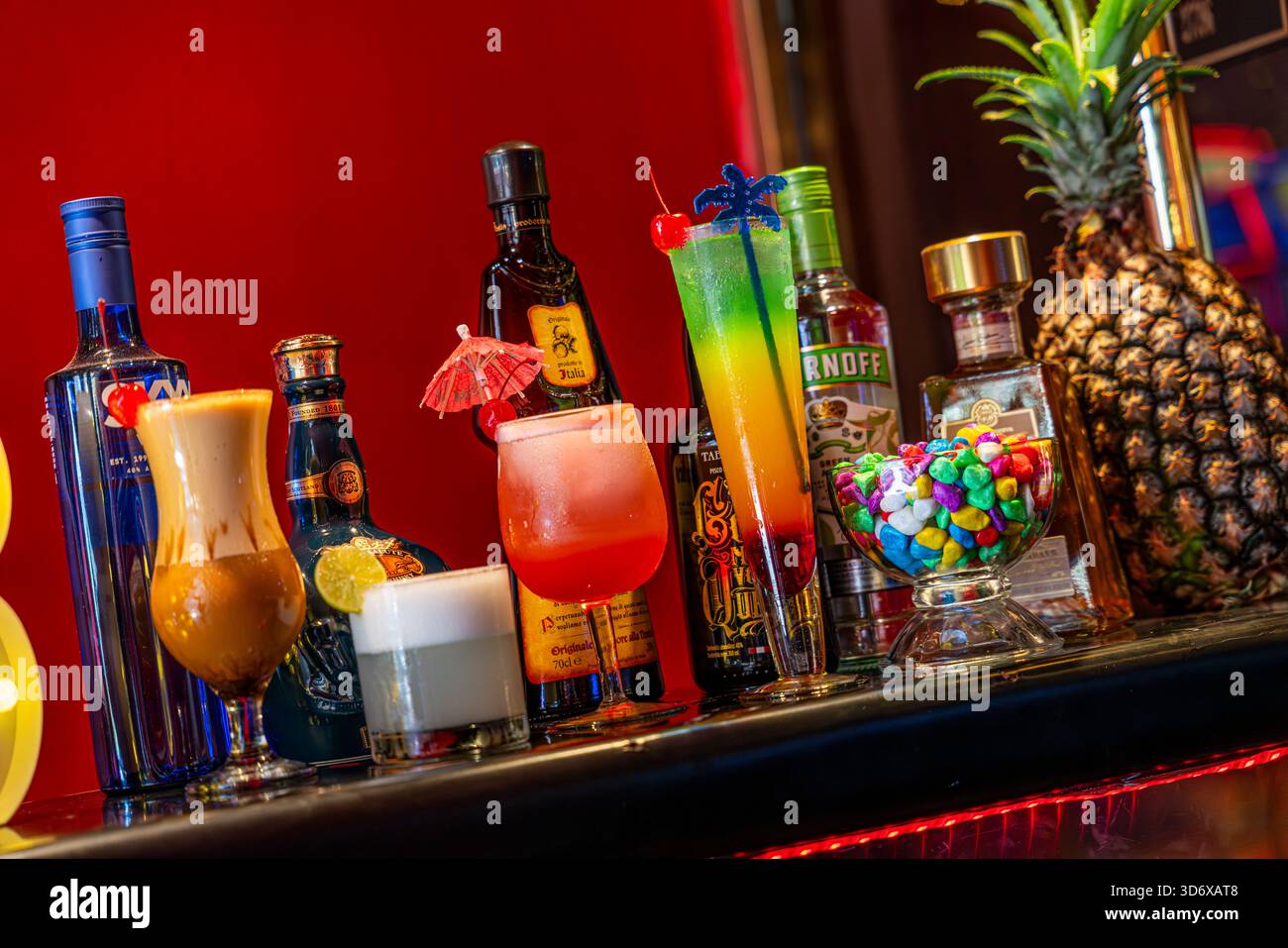 Farbenfrohe Cocktails, Spirituosenflaschen, Schüssel und dekorative Elemente auf einem festlichen Bartisch mit leuchtend roter Hintergrundbeleuchtung. Stockfoto