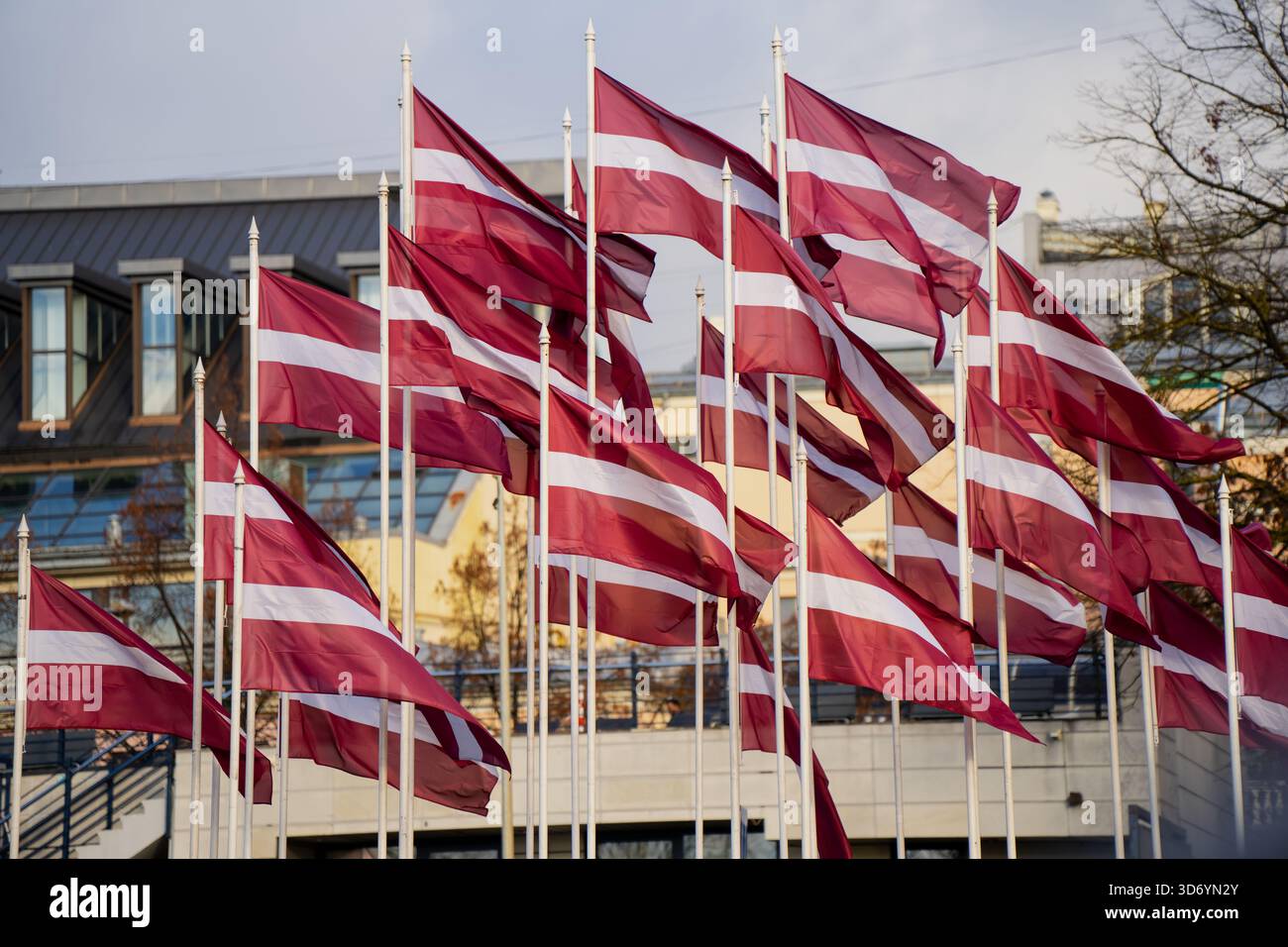 Eine Gruppe lettischer Nationalflaggen, die auf hohen weißen Pfählen montiert sind, die auf einem urbanen Außenplatz zusammenwinken, mit modernen Bauelementen, die starke Kräfte erzeugen Stockfoto