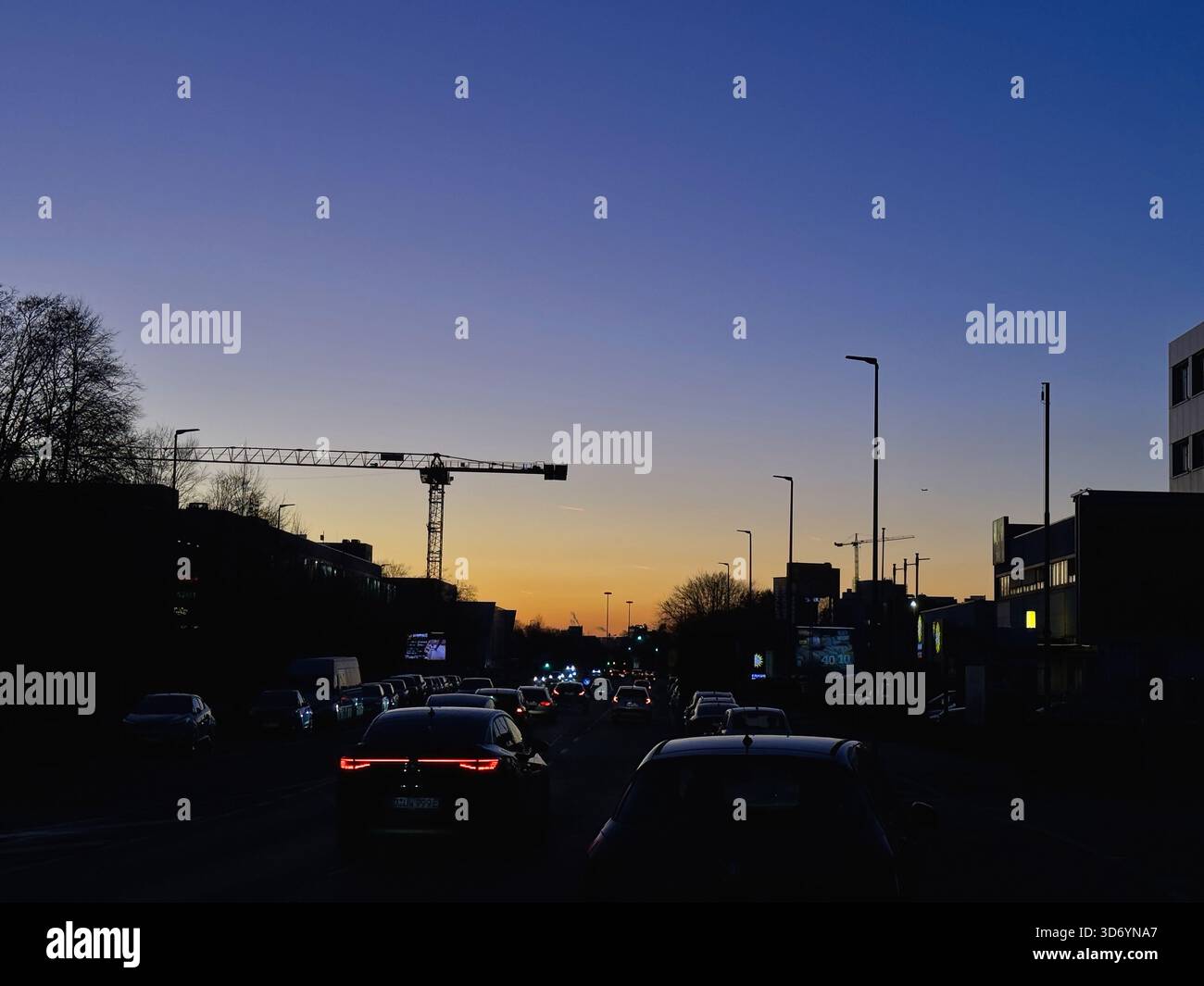 In Neuss, Deutschland, fließt der Verkehr in der Dämmerung entlang der Straße. Ein farbenfroher Sonnenuntergang erleuchtet den Himmel, während ein Baukran über den Gebäuden thront Stockfoto