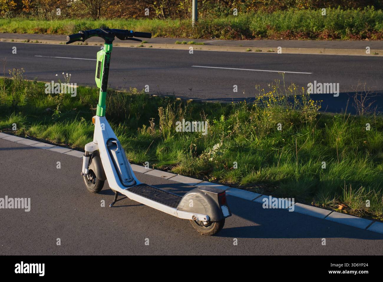 Ein grün-weißer Elektroroller liegt am Straßenrand in Neuss. Die Sonne beleuchtet die Szene und zeigt das Herbstlaub in der Nähe. Stockfoto