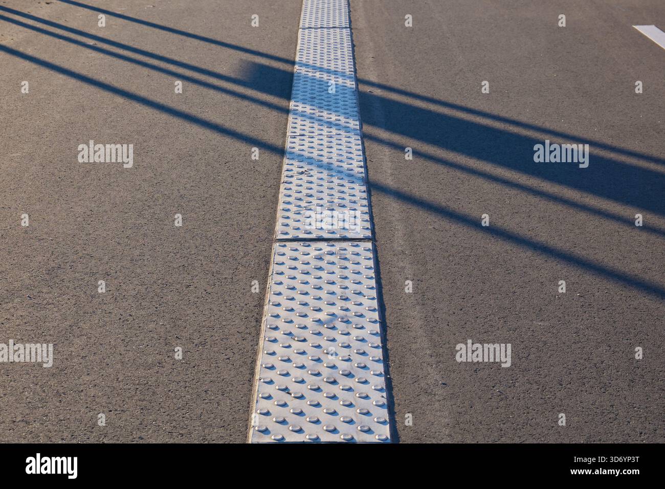 Entlang der Straße verläuft ein taktiler Metallpflasterstreifen, der sehbehinderten Personen Orientierung bietet. Die Sonne wirft Schatten auf die strukturierte Oberfläche Stockfoto