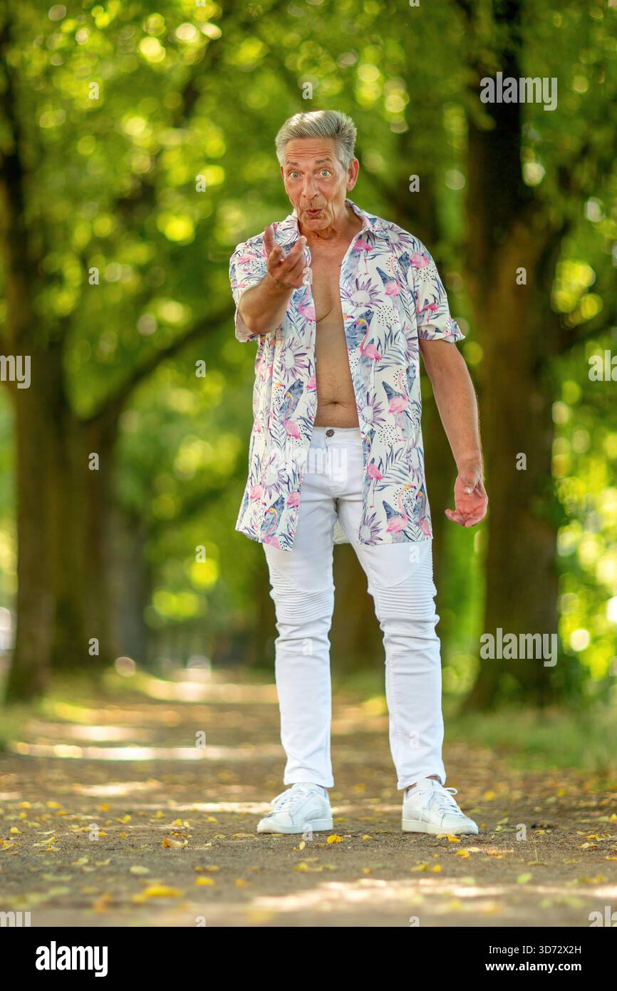 Ein fröhlicher Seniorenmann, der auf einem von Bäumen gesäumten Weg steht, ein farbenfrohes tropisches Hemd und weiße Jeans trägt. Er bewegt sich spielerisch zur Kamera und strahlt an einem sonnigen Tag in einer parkähnlichen Umgebung Selbstvertrauen und Humor aus. || Modell freigegeben Stockfoto
