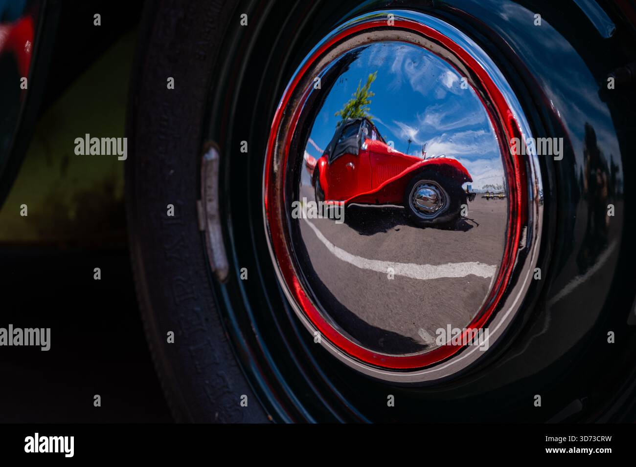 Nahaufnahme einer polierten, verchromten Radkappe, die einen knallroten Oldtimer unter einem sonnigen blauen Himmel reflektiert. Stockfoto